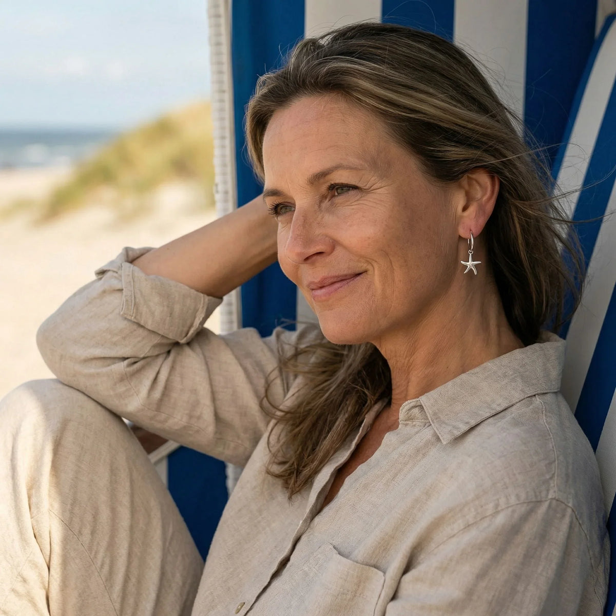 Frau mit silbernem Seestern-Ohrring sitzt entspannt am Strand in maritimer Kulisse