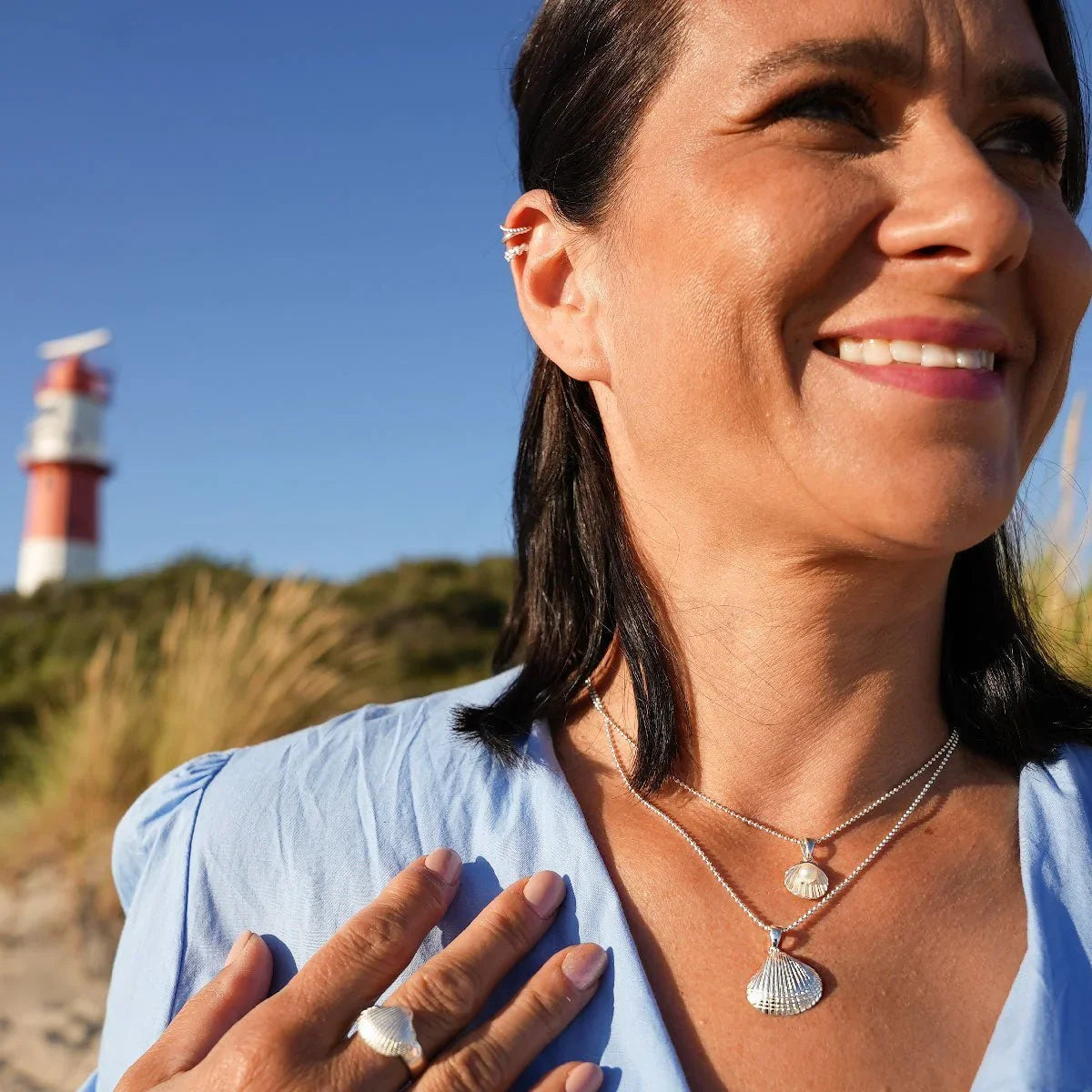 Frau mit silberner Herzmuschel-Kette und Ring am Strand vor Leuchtturm