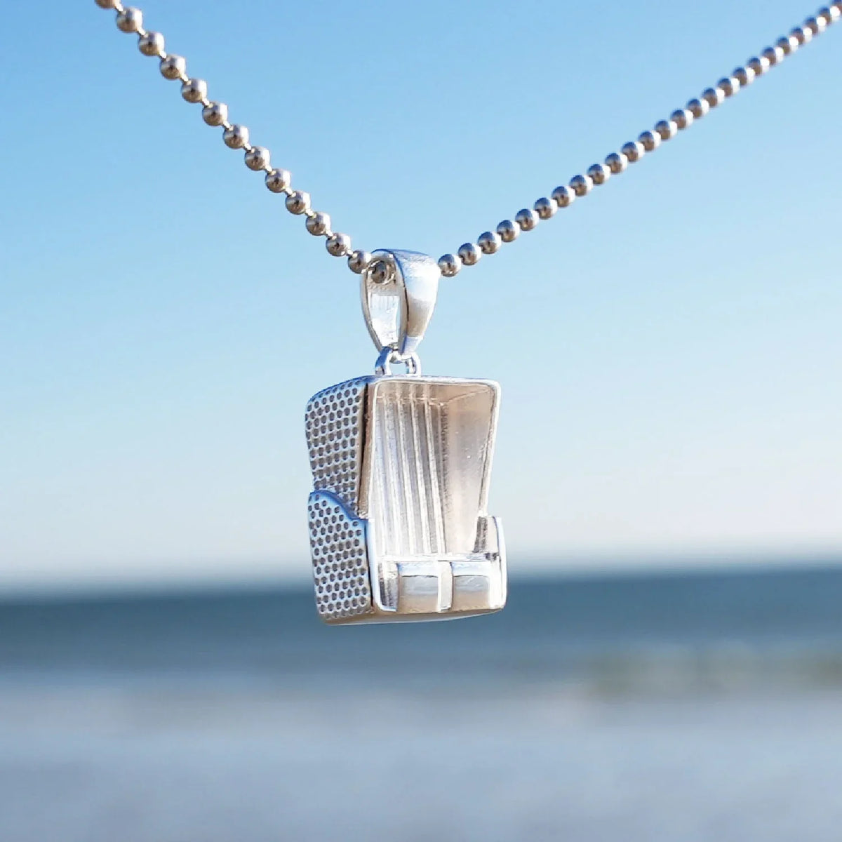 Silberner Strandkorb-Anhänger an Kette vor blauem Himmel und Meer, maritimer Schmuck