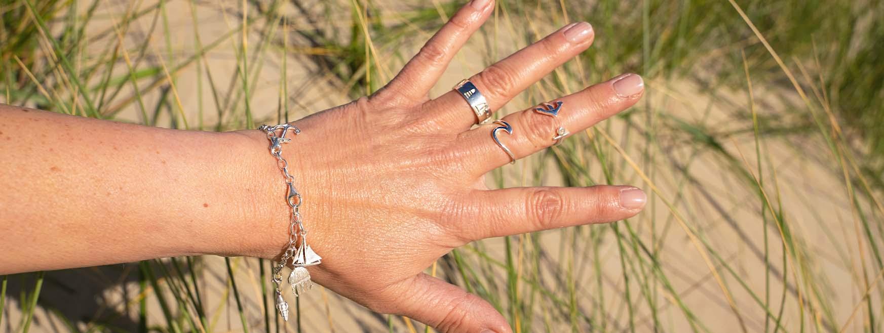 Hand mit maritimen Silberringen und Armband mit Anhängern am Strand im Sand und Dünengras