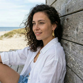 Frau mit goldenen Miesmuschel-Creolen-Ohrringen am Strand vor Holzwand