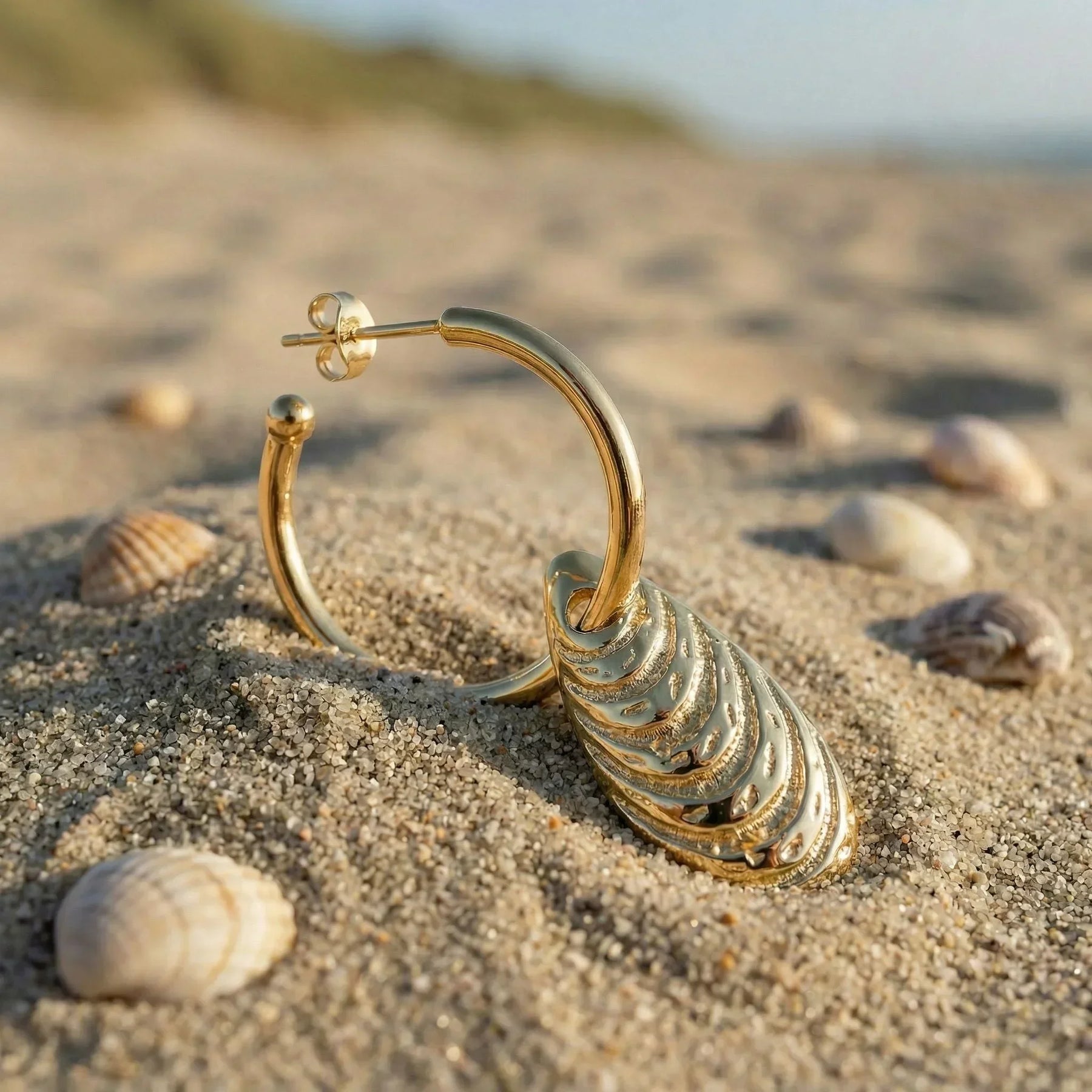 Goldener Creolen-Ohrring mit Muschelanhänger liegt im Sand am Strand, umgeben von Muscheln