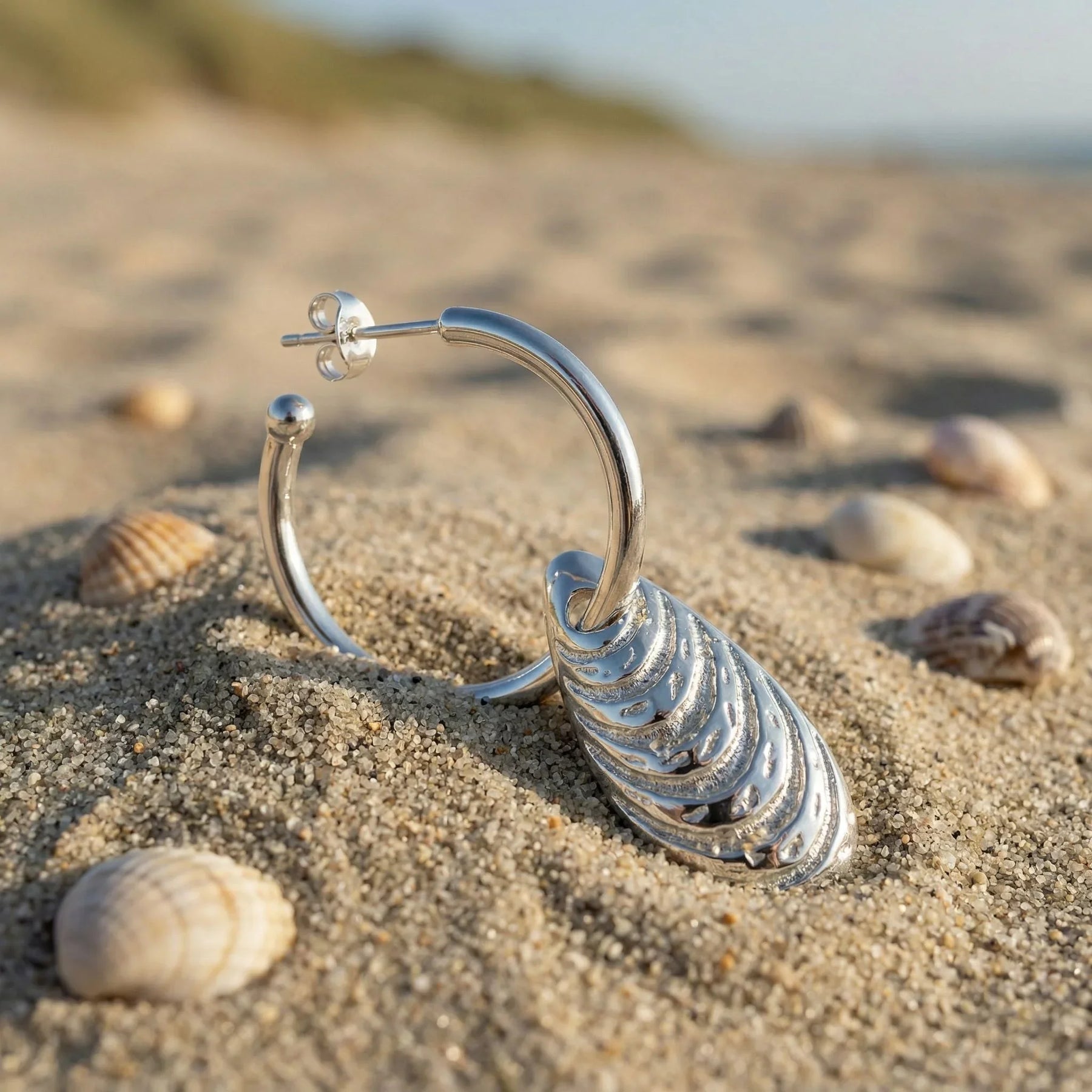 Creolen-Ohrring mit silbernem Miesmuschel-Anhänger am Sandstrand zwischen Muscheln