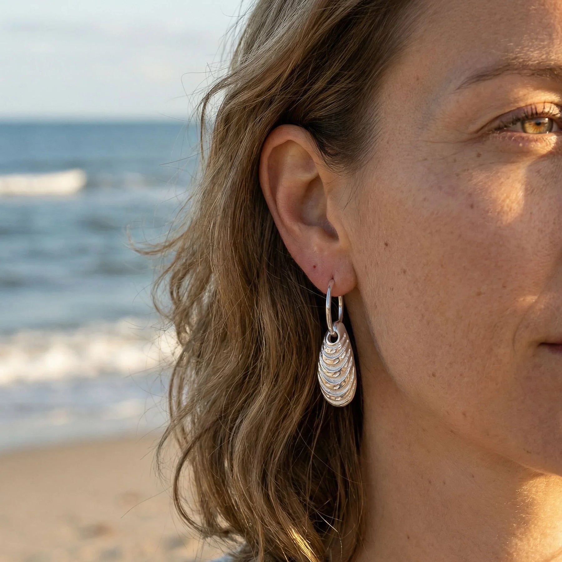Silberner Miesmuschel-Ohrring am Ohr einer Frau am Strand, maritimer Schmuck