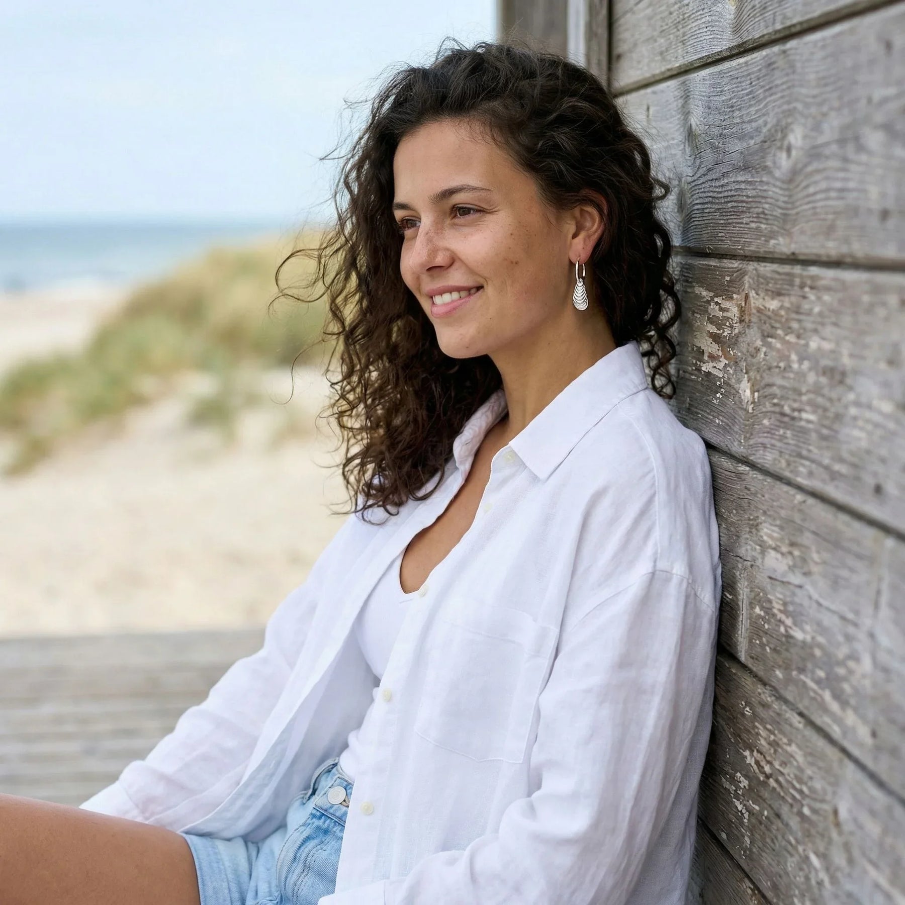 Frau am Strand mit silbernen Creolen-Ohrringen in Muschelform, maritime Schmuckmode