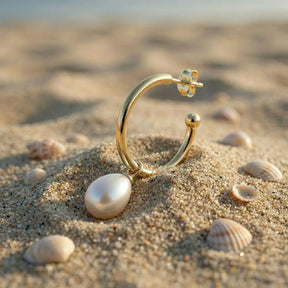 Goldener Creolen-Ohrring mit Perlenanhänger im Sand am Strand, Muscheln im Hintergrund