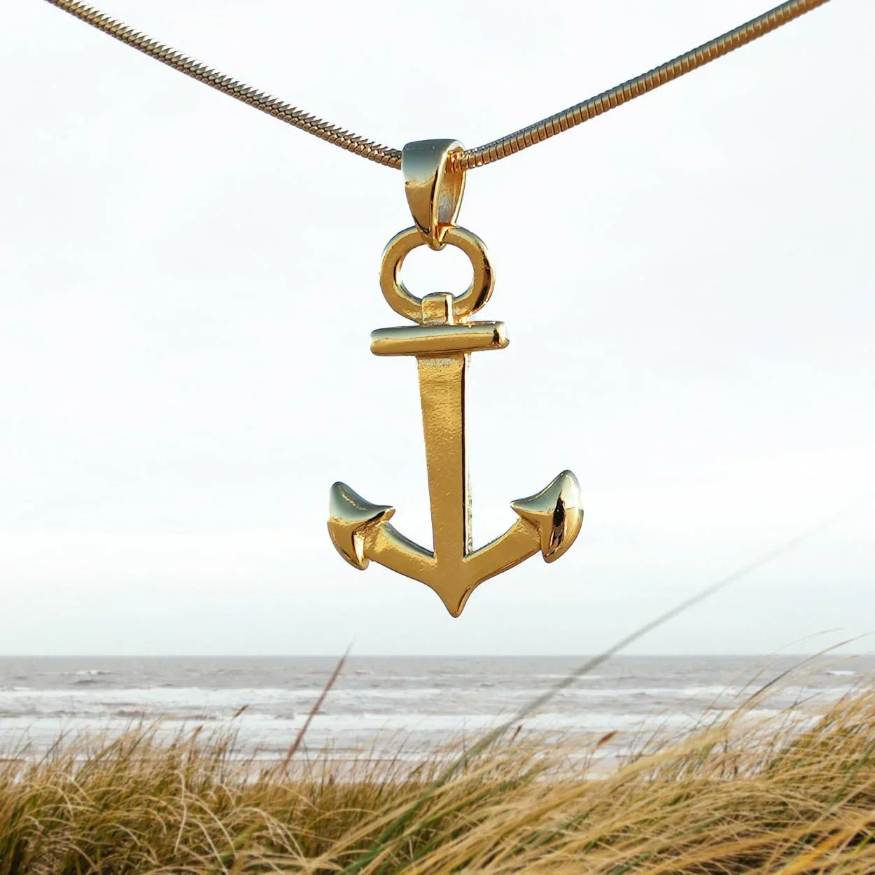 Goldener Anker-Anhänger an Kette vor Strand und Meer, maritimer Schmuck