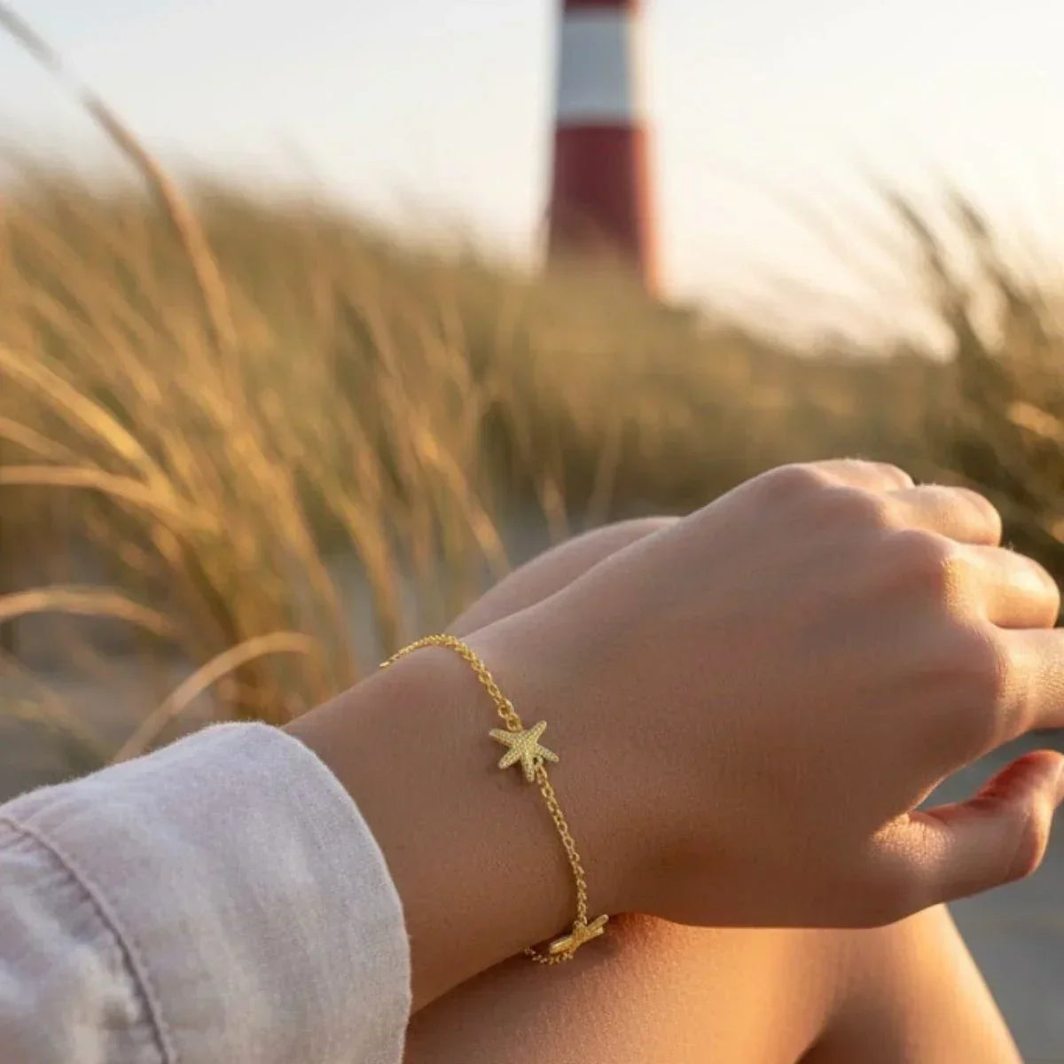 Goldenes Armband mit Seestern-Anhänger am Handgelenk, maritimer Schmuck am Strand vor Leuchtturm