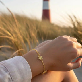 Goldenes Armband mit Seestern-Anhänger am Handgelenk, maritimer Schmuck am Strand vor Leuchtturm