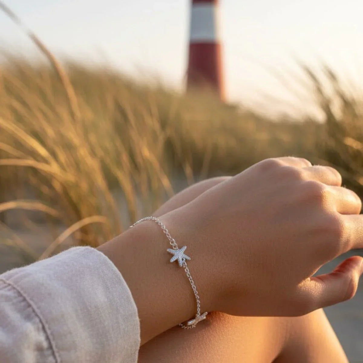 Silber Armband mit Seestern-Anhängern am Handgelenk, maritimes Motiv, Leuchtturm im Hintergrund