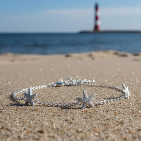 Silbernes Armband mit Seestern-Anhängern auf Sandstrand, Meer und Leuchtturm im Hintergrund