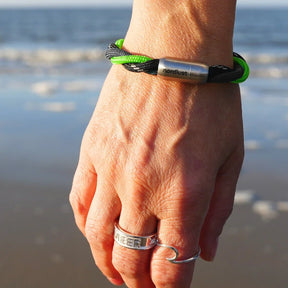 Hand mit maritimem Armband aus schwarzem und grünem Seil am Strand, Meer im Hintergrund