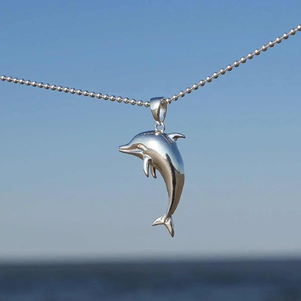 Silberner Delfin-Anhänger an Kette vor blauem Himmel und Meer, maritimer Schmuck