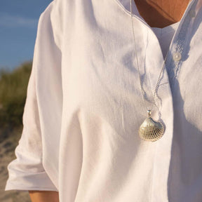 Silberne Herzmuschel-Anhänger-Kette auf weißer Bluse am Strand bei Sonnenlicht