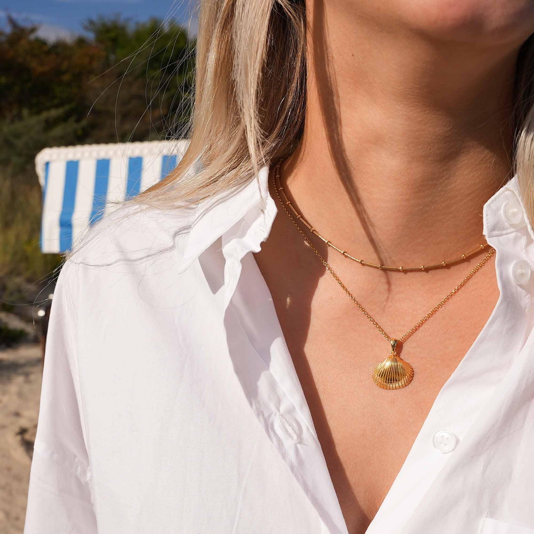 Frau mit goldener Herzmuschel-Kette am Strand, weißes Hemd, maritimer Schmuck