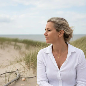 Frau mit goldener Herzmuschel-Kette am Strand mit Dünen und Meer im Hintergrund