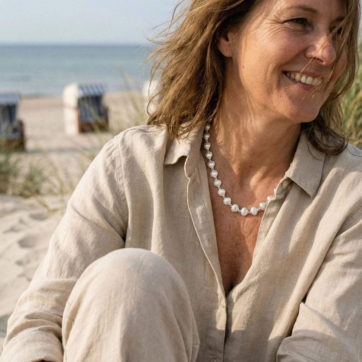 Frau mit beige Leinenhemd und maritimer Muschelkette am Strand, Meer im Hintergrund