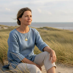 Frau mit goldener Muschelkette sitzt entspannt in Dünen am Meer, maritimer Schmuck, Strand