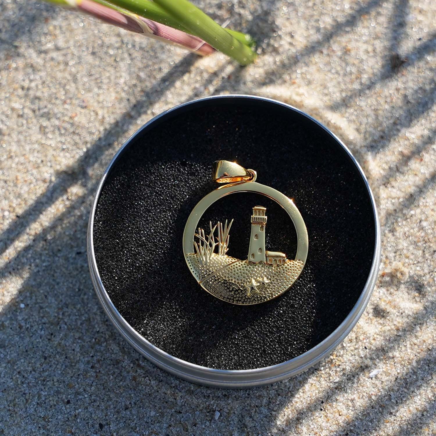 Goldener Anhänger mit Leuchtturm-Motiv im maritimen Stil auf Sand in Schmuckdose