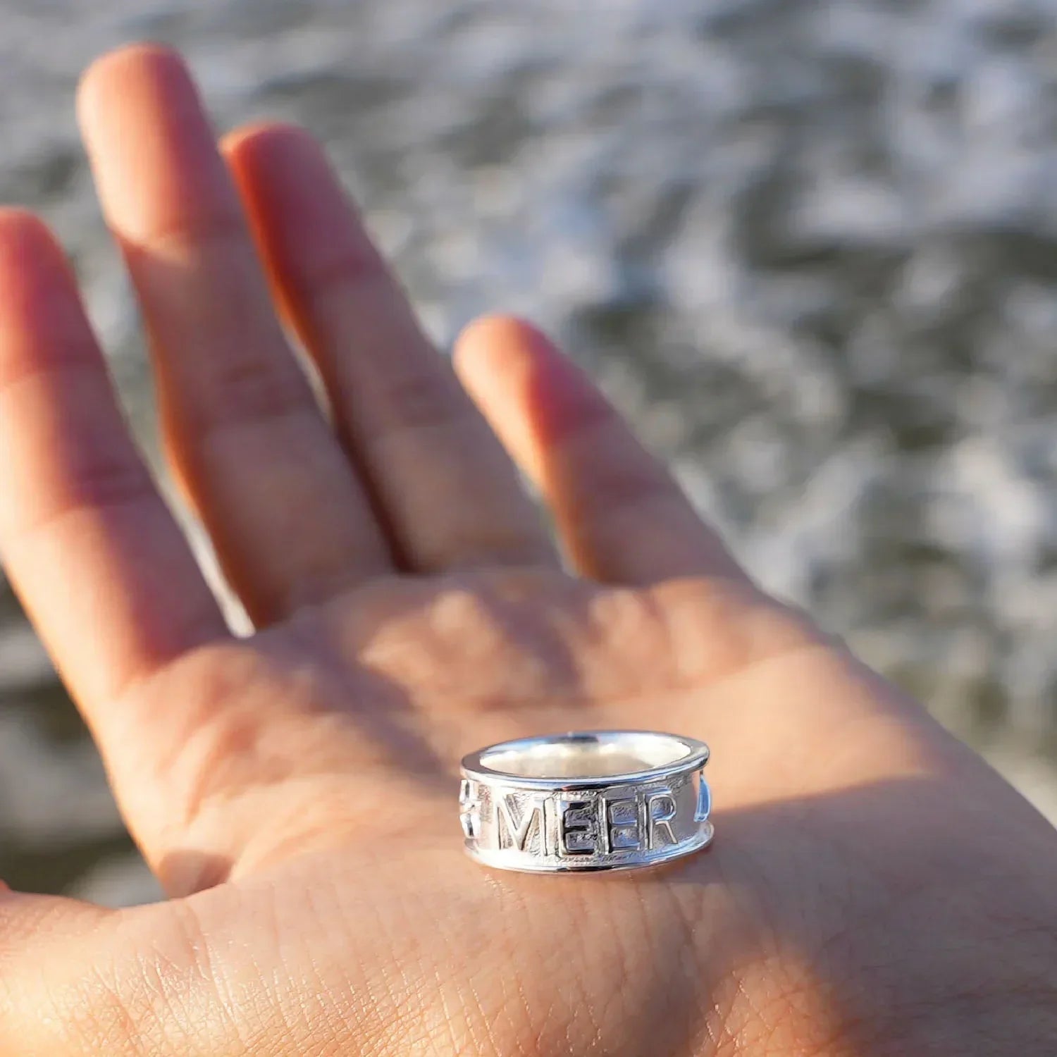 Silberner Ring mit Gravur 'Meer' auf einer Hand am Strand, maritimer Schmuck