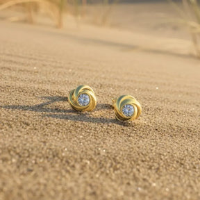 Goldene Ohrstecker mit Knotendesign und funkelndem Stein auf Sand am Strand