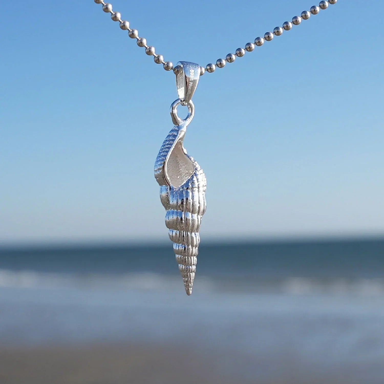 Silberner Muschel-Anhänger an Kette, maritimer Schmuck vor Meer und blauem Himmel