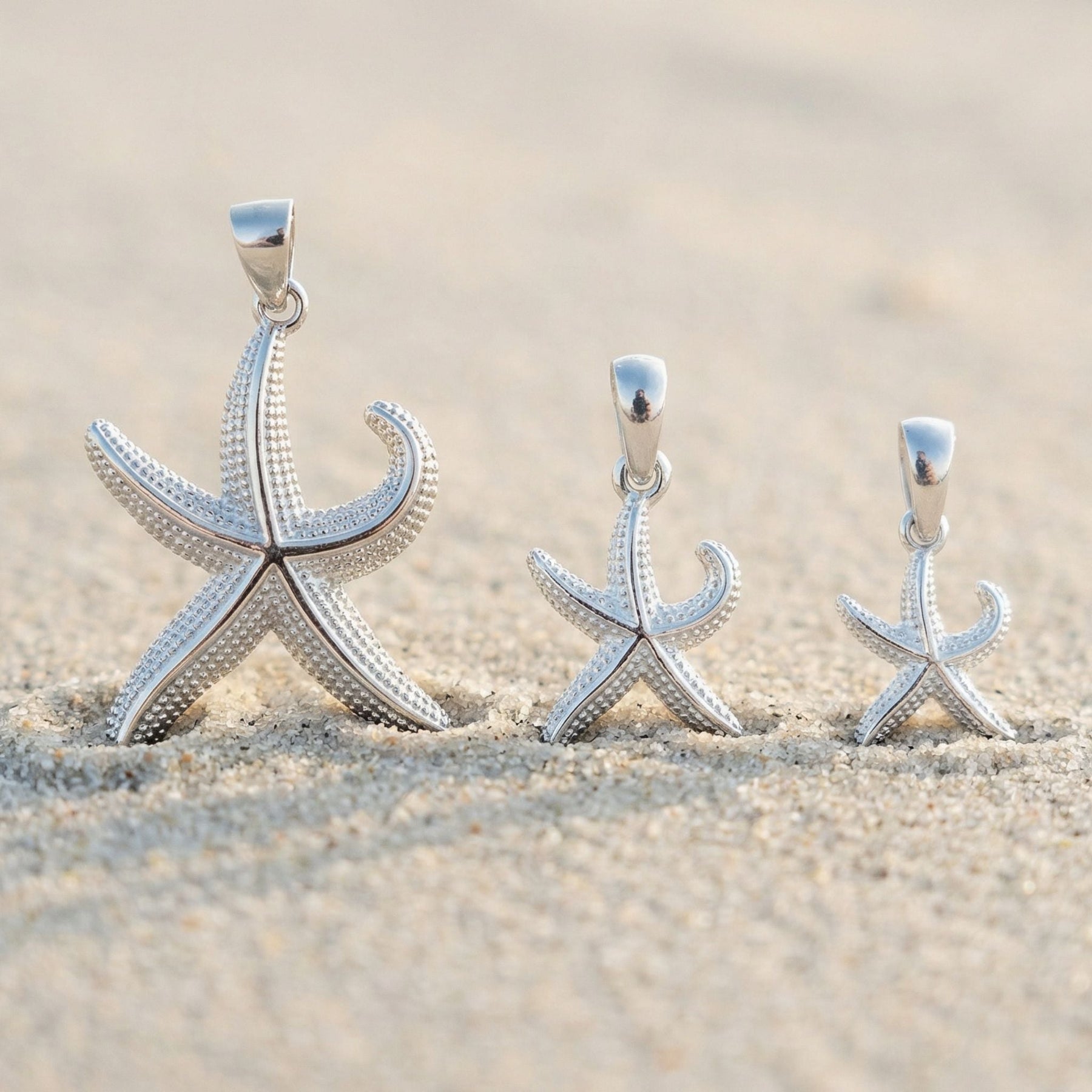 Drei silberne Seestern-Anhänger in verschiedenen Größen im Sand am Strand