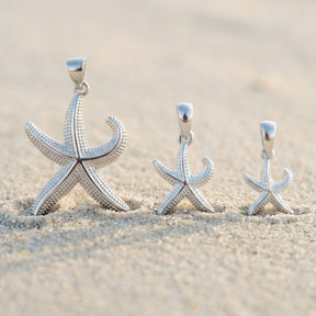 Drei silberne Seestern-Anhänger in verschiedenen Größen im Sand am Strand