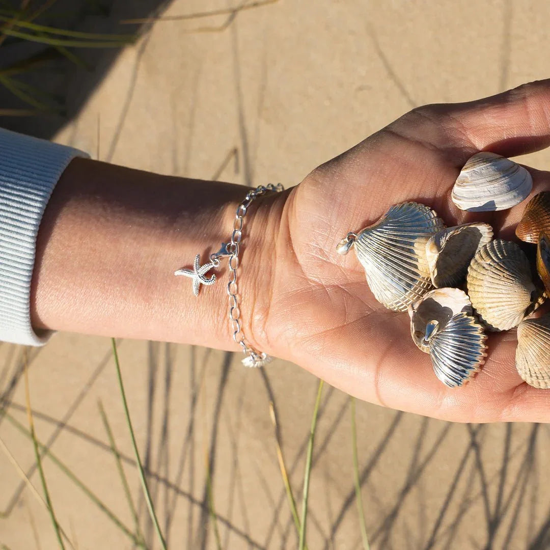 Silbernes Seestern-Armband am Handgelenk, Muscheln in der Hand, am Strand