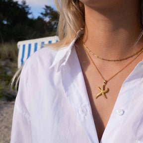 Frau am Strand mit goldener Seestern-Kette und weißer Bluse, maritimer Schmuck