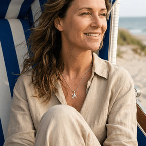 Frau mit silberner Seestern-Kette sitzt entspannt am Strand in maritimer Umgebung.