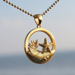 Goldener Anhänger mit Seestern-Motiv, maritimer Kette vor unscharfem Strandhintergrund