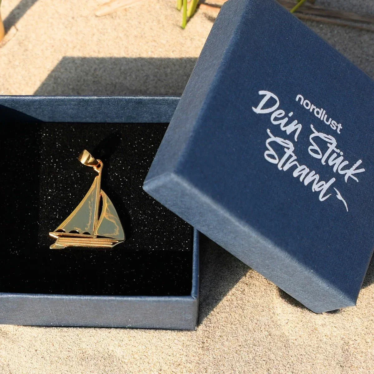 Goldener Segelboot Anhänger in Geschenkbox auf Sand, maritimer Schmuck