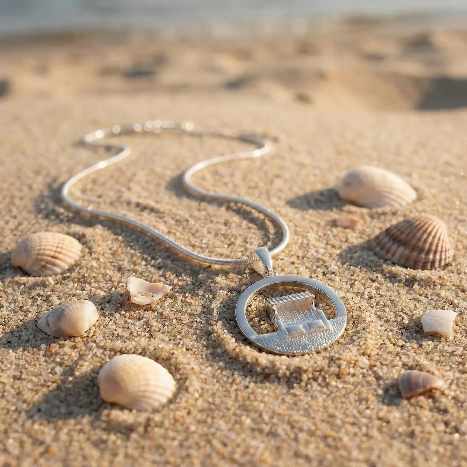 Silberkette mit Strandkorb-Anhänger im Sand, umgeben von Muscheln am Strand