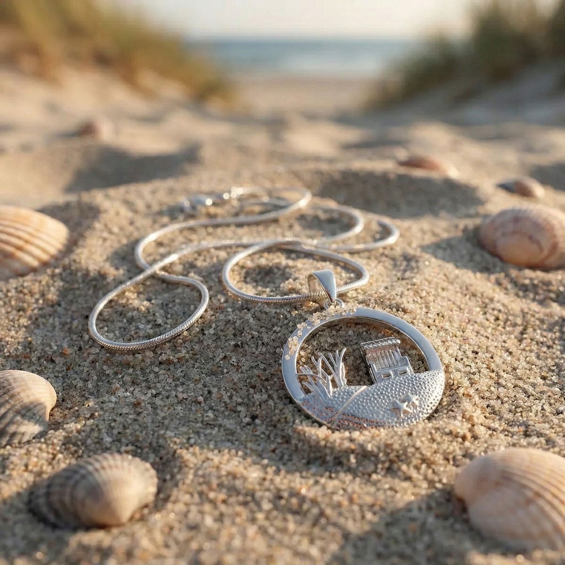 Silberne Strandkorb-Kette im Sand am Meer mit Muscheln, maritimer Schmuck, Küstenmotiv
